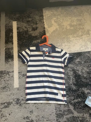 Randig pikétröja från Abercrombie & Fitch - Snygg pikétröja med marinblå och vita ränder från Abercrombie & Fitch. Klassisk krage, korta ärmar och liten logga på bröstet. Perfekt till jeans eller shorts för en avslappnad look.