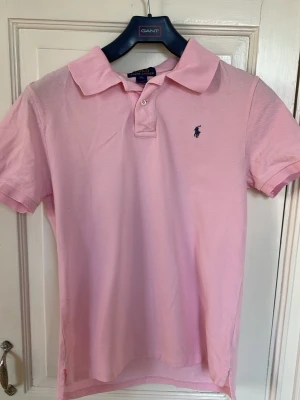 Rosa pikétröja från Polo Ralph Lauren - Snygg rosa pikétröja från Polo Ralph Lauren med klassisk krage och två knappar framtill. Tröjan har korta ärmar och den ikoniska broderade loggan på bröstet. Tillverkad i mjuk bomull som känns skön mot huden.