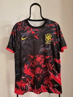 Nike Brasil svart-röd tränings-tshirt - Snygg svart och röd tränings-tshirt från Nike med Brasil-logga och gult swoosh på bröstet. Framsidan har ett stort tigertryck och baksidan har röda rivmärken. Tillverkad i lätt och ventilerande material, perfekt för träning eller match.