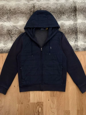 Ralph Lauren Cardigan/Hybrid Zip Up Hoodie - Hej! 👋 Säljer en sjukt snygg Marinblå Polo Ralph Lauren Hybrid Zip Hoodie/Cardigan i storlek M. En riktig premiummodell med stickade ärmar/rygg och ett quiltat, vindsäkert framparti 🧥 Mycket gott skick! Det kan förekomma någon liten tråd som sticker upp, men inget som stör eller påverkar utseendet ✨ Sitter perfekt (Modell: 177cm) och har den klassiska pony-loggan broderad i grått på bröstet 🐎 Detta är det perfekta lyxiga basplagget till garderoben. Fraktar snabbt! 🚛📦💬💯 