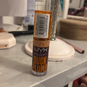 NYX Duck Plump läppglans orange - NYX Duck Plump läppglans med transparent orange lock och tydlig logga. Ger en plumpande effekt och har en intensiv, glansig finish. Förpackningen är kompakt och enkel att ta med i väskan. Perfekt för dig som vill ha fylligare läppar och en trendig look.