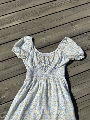 Blommig sommarklänning med puffärm - Söt klänning med ljusblått och gult blommönster, puffärmar och resår i midjan. Klänningen har en rundad halsringning och korta ärmar med resår. Den har även en fin liten rosett framtill!