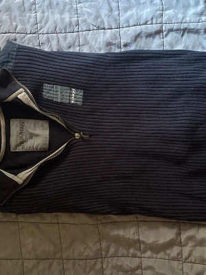 Mörkblå ribbad tröja från Sebago - Mörkblå ribbstickad tröja från Sebago med halv dragkedja och ståkrage. Tröjan har diskret Sebago-logga på bröstet och kontrastfärgade detaljer vid kragen. Perfekt för dig som gillar klassisk och stilren design.