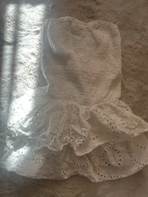 Vit axelbandslös kortklänning från meet me there - Söt vit axelbandslös kortklänning med smockad överdel och volangig kjol i broderad bomull. Perfekt för varma dagar och har en romantisk känsla med sina detaljerade broderier och luftiga lager.