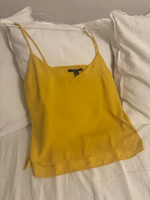 Gul linnetopp från Forever 21 - Snygg gul linnetopp från forever 21 med tunna axelband och v-ringad hals. Toppen har en lös passform och är tillverkad i ett mjukt, lent material som känns skönt mot huden. Perfekt för varma dagar och enkel att styla med jeans eller kjol.