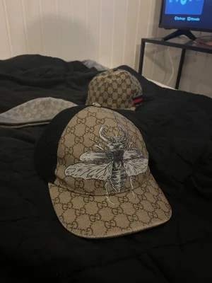 Beige gucci keps - Säljer en snygg Gucci keps i storlek M med klassiskt beige GG-mönster och stor bi-illustration framtill. Kepsen har svart mesh på sidorna och justerbart band bak med röd-gröna detaljer. Materialet är en mix av bomull och polyester.