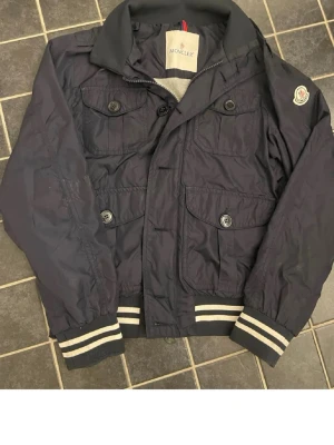 Mörkblå bomberjacka från Moncler - Snygg mörkblå bomberjacka från Moncler med fyra framfickor och vita ränder på muddar och nederkant. Jackan har en klassisk krage, knappstängning och Moncler-logga på ärmen. Tillverkad i ett glansigt, lätt material som ger en sportig look.