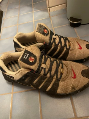 Nike Shox beige och svarta sneakers - Nike Shox sneakers i beige och svart med röda detaljer och guldaccenter. Skorna har klassisk Shox-dämpning, mesh och syntetmaterial, samt randigt innerfoder och tydliga Nike-loggor på plös och sula. Snygg retrostil för dig som gillar streetwear. Är lite slitna men säljer för 50 procent mindre av vad den är värd, pallar inte fixa skorna, kostar max 100-200 att göra dem helt nya sen är värdet på dem ungefär 4000 