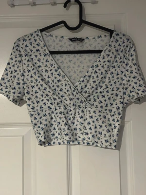 Vit blommig crop top - Söt vit crop top med blått blommönster och omlott-ringning. Korta ärmar och feminin passform perfekt till höga jeans, linnebyxor eller kjol för en somrig och trendig outfit (ifrån shein) 
