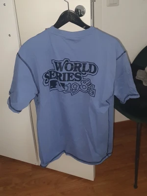 Blå World Series 1984 T-shirt - Säljer en blå T-shirt från New Era med tryck 'World Series 1984' på ryggen och ett stort D på bröstet. T-shirten har kontrastsömmar och klassisk passform. Perfekt för dig som gillar sportig stil och retrovibbar.
