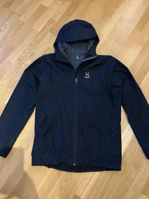 Svart vindjacka från Haglöfs - Snygg svart vindjacka från Haglöfs med huva och hel dragkedja framtill. Jackan har två snedställda fickor med dragkedja och diskret logga på bröstet. Perfekt för blåsiga dagar och enkel att matcha med olika outfits.