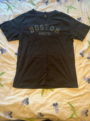 Svart Boston South t-shirt - Svart t-shirt från SDC med trycket 'BOSTON SOUTH' på bröstet. Klassisk rund hals och korta ärmar. Perfekt för en avslappnad och sportig stil. Tillverkad i mjukt bomullsmaterial.