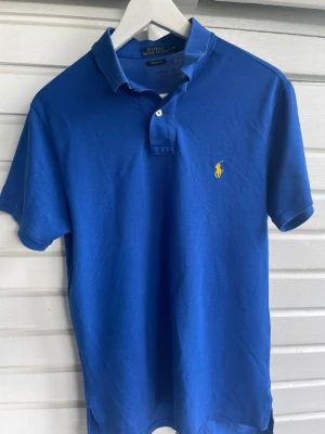 Blå pikétröja från Polo Ralph Lauren - Klassisk blå pikétröja från Polo Ralph Lauren med gul broderad logga på bröstet. Tröjan har korta ärmar, krage och knappar vid halsen. Tillverkad i 100% bomull för en skön och luftig känsla.