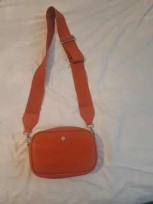 Orange axelväska  - Snygg orange axelväska med justerbar bred rem och silverfärgad knappdetalj framtill. 