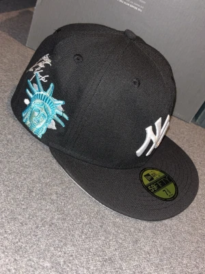Svart New Era NY keps med brodyr - Säljer en svart New Era 59FIFTY keps med vit NY-logga framtill och broderad frihetsgudinna i turkos och vitt på sidan. Texten 'Gateway to New York' är broderad ovanför motivet. Klassisk flat brim och strukturerad passform.
