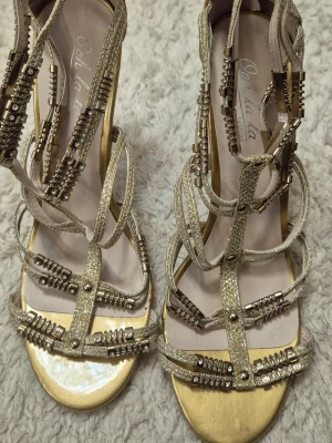 Guldiga sandaler från Oh la la fashion - Säljer ett par glittriga guldiga sandaler från Oh la la fashion. Sandalerna har flera tunna remmar dekorerade med metallpärlor och nitar, samt öppen tå. Perfekta för dig som vill sticka ut med en glamorös look.