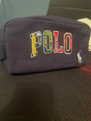 Necessär Polo Ralph Lauren marinblå - Helt ny Necessär aldrig använd, nypris 1200kr. i marinblått tyg från Polo Ralph Lauren med broderad POLO-text i flera färger och klassisk vit logga. Rektangulär form med dragkedja och innerfoder i matchande blått. Perfekt för att samla småprylar. 