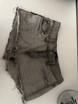 Gråa jeansshorts med råa kanter - Säljer ett par gråa jeansshorts med råa, fransiga kanter och slitna detaljer. Shortsen har klassiska fickor fram och bak, bälteshällor och knappstängning. Perfekta för en avslappnad och edgy stil.
