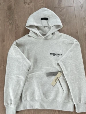 Grå Essentials hoodie med huva - Snygg ljusgrå hoodie från Essentials med svart logga på bröstet och stor känguruficka framtill. Klassisk modell med huva och ribbade muddar vid ärmar och nederkant. Perfekt för en avslappnad streetwear-look.