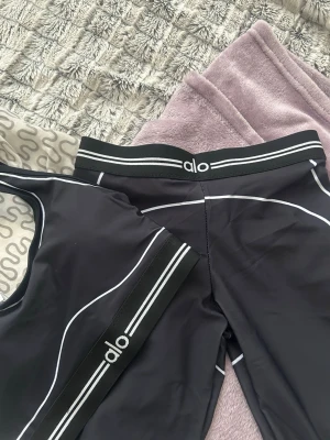 Svart träningsset från Alo - Snyggt svart träningsset från Alo med vita kontrastsömmar och sportiga ränder. Setet består av en sport-bh och tights, båda med elastiska band och Alo-logga. Perfekt för gymmet eller yoga. Materialet är stretchigt och följsamt.
