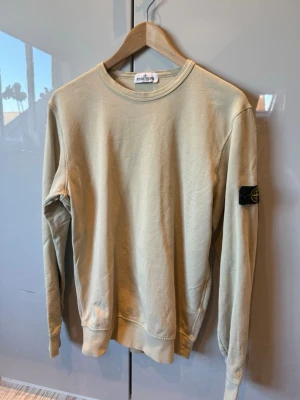 Beige sweatshirt från Stone Island - Säljer en beige sweatshirt från Stone Island med klassisk rund hals och ribbade muddar. Tröjan har den ikoniska svarta patchen med gul logga på vänster ärm. Perfekt för dig som gillar streetwear och enkel stil.