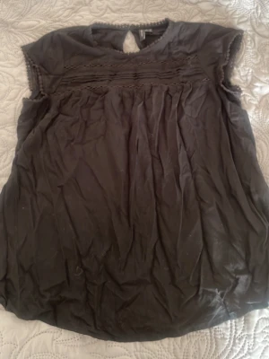 Svart blus från Vero Moda - Svart blus från Vero Moda med rund halsringning och dekorativ knapp i nacken. Blusen har korta ärmar med små bollar längs kanten och fina veck framtill. Tillverkad i ett mjukt, tunt tyg som ger ett avslappnat intryck.