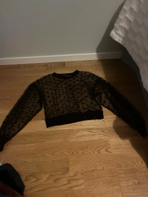 Svart mesh topp med prickigt mönster - Trendig svart mesh topp med lång ärm och prickigt mönster. Croppad modell med ribbade muddar vid ärmslut och midja. Perfekt för att addera en edgy vibe till din outfit.