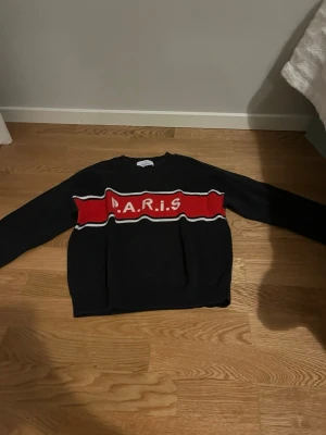 Svart tröja med röd PARIS-text - Cool svart tröja med en bred röd rand över bröstet där det står 'P.A.R.I.S' i vita bokstäver. Tröjan har rund halsringning och långa ärmar. Perfekt för dig som gillar streetwear och vill sticka ut lite extra.