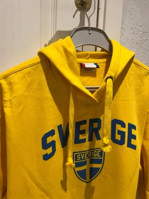 gul huvtröja, storlek XL - Gul huvtröja med trycket "SVERIGE" i blått på framsidan. Tröjan har en känguruficka framtill och muddar vid ärmslut och nederkant.