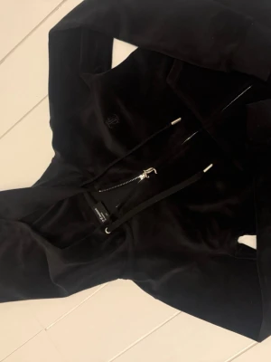 Svart velourset från Juicy Couture - Säljer ett svart set från Juicy Couture med zip-hoodie och matchande byxor. Hoodien har dragkedja och broderad logga på bröstet, byxorna har resår i midjan och raka ben. Perfekt för en avslappnad och trendig look. Dock är de storlek xxs i byxorna och i tröjan xs. På ett av snörena på byxorna har den silvriga detaljen lossnat.