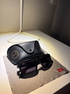 Ray-Ban Justin Solglasögon - Säljer ett par snygga svarta Ray-Ban solglasögon med breda bågar och mörka glas. Klassisk fyrkantig form och Ray-Ban-logga på sidan. Medföljer originalfodral och putsduk. Perfekt för dig som vill ha en ikonisk och stilren look. 🎉Glasögonen har en liten repa (se bild 5) på bågen. 🎉 Glasögonen köpte i Synsams butik i 2024 för 1695kr, säljs på grund av nya köpta och repa! 🎉För mer info skriv dm!
