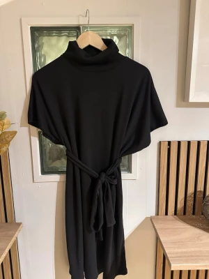 Svart ribbad klänning med polokrage - Svart ribbad klänning med polokrage och knytskärp i midjan. Klänningen har korta ärmar och en loose passform som ger en avslappnad men stilren look. Perfekt för dig som gillar minimalistisk stil och vill ha något bekvämt men snyggt.