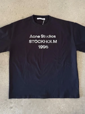 Svart Acne Studios t-shirt - Svart t-shirt från Acne Studios med vit text 'Acne Studios STOCKHOLM 1996' på bröstet. Klassisk rund halsringning och rak passform. T-shirten är på storlek M och är perfekt för dig som gillar stilrena och trendiga plagg med en touch av streetwear.