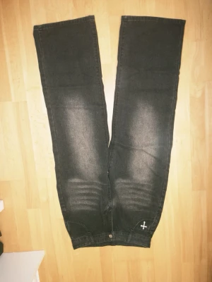 Svarta raka jeans med korsdetaljer - Säljer ett par svarta jeans med raka ben och coola korsdetaljer på bakfickorna samt ett litet kors framtill. Jeansen har en tvättad look och är tillverkade i denim. Perfekta för dig som gillar alternativ stil.