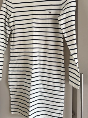 Randig långärmad klänning Gant - Klassisk vit och marinblå randig klänning från Gant med lång ärm och rund hals. Klänningen har broderad logga på bröstet och är gjord i mjuk bomull. Perfekt för en avslappnad och stilren look.