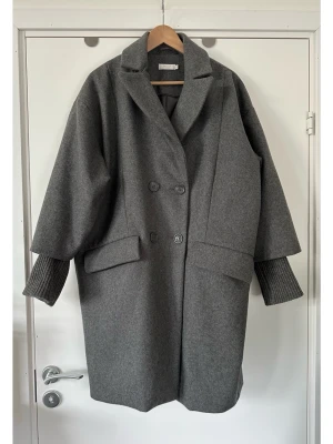 WERA - Grå oversized kappa - storlek 40 - Stilren kappa i en tidlös grå nyans med dubbelknäppning framtill från WERA Stockholm i storlek 40. Kappan har en klassisk krage och slag, samt två fickor fram. Ärmarna är designade med en bredare passform och avslutas med en ribbad mudd, vilket ger en modern touch. En slits baktill säkerställer god rörelsefrihet. 65% bomull, så bra material som andas. Nypris 1899:-. Mycket gott skick, som ny!