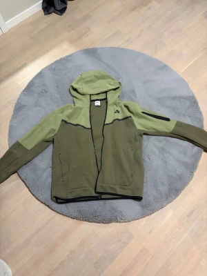 Grön hoodie från Nike med dragkedja - Snygg tvåfärgad hoodie från Nike i olivgrönt och ljusgrönt. Modellen har hel dragkedja, huva och svarta detaljer. Ficka på ärmen och två sidofickor. Perfekt för en sportig och avslappnad stil.