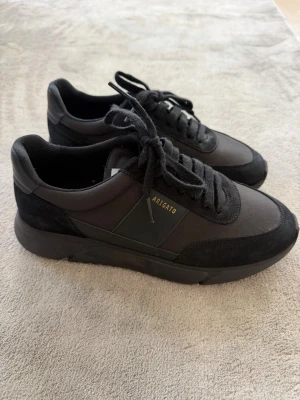 Svarta sneakers från Axel Arigato - Snygga svarta sneakers modell ” Genesis Vintage Runners”från Axel Arigato med stilren design. Skorna har ovandel i textil och mocka, svarta skosnören och diskret logga i guld på sidan. Perfekta för dig som gillar minimalistisk och modern stil.