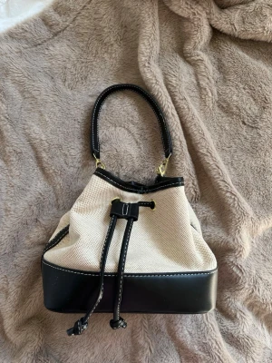 Beige och svart bucket bag - Stilren bucket bag i beige canvas med svarta detaljer, helt oanvänd och i nyskick, man får med ett kort och ett långt band