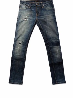 Nudie jeans thight long john - Säljer ett par blå jeans från Nudie Jeans med snygga slitningar och lagade detaljer på benen. Klassisk femficksmodell med orange sömmar och Nudie-logga på bakfickan. Jeansen har rak passform och är tillverkade i jeansmaterial med dragkedja.