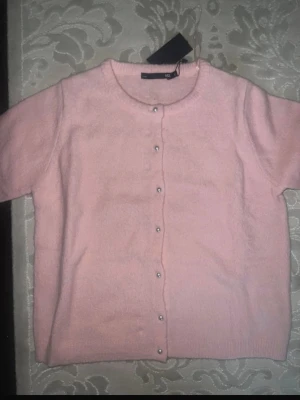 Rosa kofta med pärlknappar 157 - Söt ljusrosa kofta från 157 med korta ärmar och pärlknappar framtill. Koftan har rund halsringning och är stickad i ett mjukt material. Perfekt för att addera en gullig touch till din outfit.