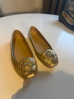 Guldiga ballerinaskor Michael Kors - Snygga ballerinaskor i metallic guld från Michael Kors med rund tå och stort MK-emblem framtill. Skorna är i glansigt läder och har en klassisk, stilren design som passar perfekt till många outfits.