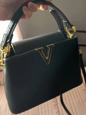 Svart handväska med gulddetaljer - Stilren svart handväska från Valentino med elegant V-logga i guld framtill. Väskan har både handtag och avtagbar axelrem, samt snygga guldfärgade metalldetaljer. Tillverkad i strukturerat läder för en lyxig känsla.