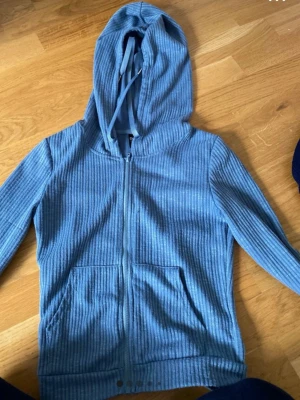 Blå ribbad hoodie med dragkedja - Säljer en blå hoodie i ribbat tyg med dragkedja framtill och kängurufickor. Tröjan har huva med dragsko och långa ärmar. Perfekt för en avslappnad och chill stil.