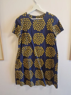 Blå klänning med gula blommor Marimekko - Superfin blå klänning med kort ärm och ett stort gult blommönster över hela plagget. Rak passform och rund halsringning, perfekt för dig som gillar färg och mönster. Tillverkad i mjuk bomull som känns skön mot huden. Handsydd i marimekkotyg