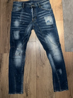 Mörkblå distressed jeans från Dsquared2 - Säljer ett par mörkblå jeans från Dsquared2 med slitna detaljer, blekta partier och färgstänk. Jeansen har klassisk femficksmodell, smal passform och synliga sömmar. Perfekta för dig som gillar en edgy och trendig stil.
