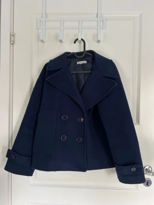 Mörkblå kappa - Mörkblå kappa från Design By Si med dubbelknäppning. Jackan har breda ärmar med knappar och två stora fickor framtill. Funkar som vårkappa och höstkappa