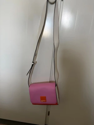 Rosa och orange axelväska Karl Lagerfeld - Cool axelväska från Karl Lagerfeld Jeans i rosa och orange med vit justerbar axelrem. Väskan har en fyrkantig form och är tillverkad i slätt konstläder. Logotypen syns på framsidan i vitt på en orange detalj. Äkta