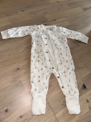 Vit pyjamas med blommönster från BeKids - Vit pyjamas från BeKids i ekologisk bomull med små blommönster i gult, rött och grått. Långärmad med knäppning framtill och fotdelar. Perfekt för mysiga kvällar och sköna nätter.
