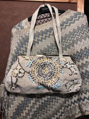 Broderad axelväska med paljetter - Axelväska i beige tyg med broderat blommönster och färgglada paljetter i blått, gult och lila. Väskan har metallspänne upptill och två matchande handtag. Perfekt för dig som gillar bohemisk och kreativ stil.